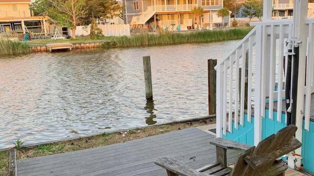 Eine Holzterrasse mit zwei Adirondack-Stühlen mit Blick auf ein Gewässer, einem weißen Zaun und einem Gebäude im Hintergrund.