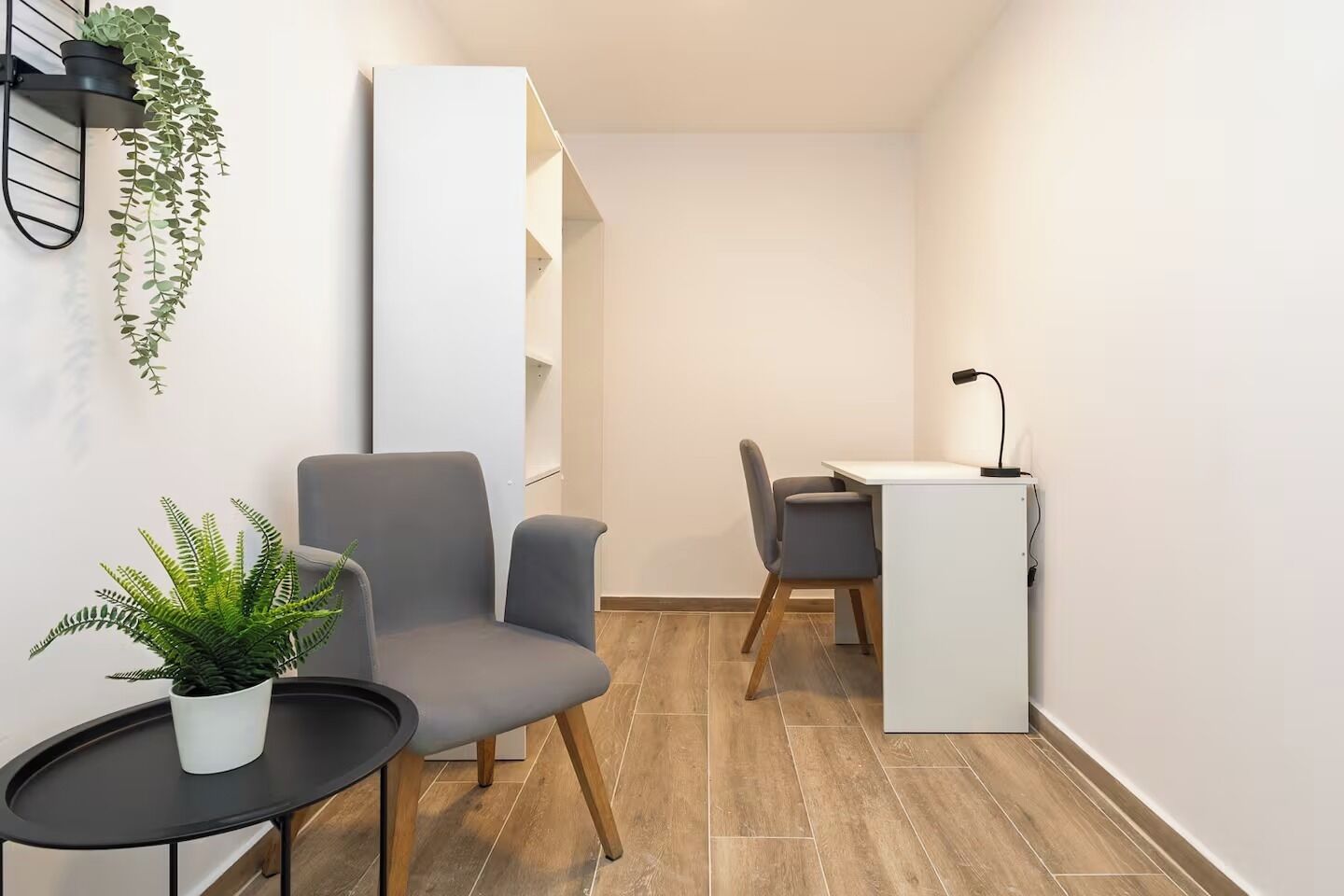 A modern room with a grey armchair, a small round table with a potted plant, a white desk with a chair, and a black lamp.