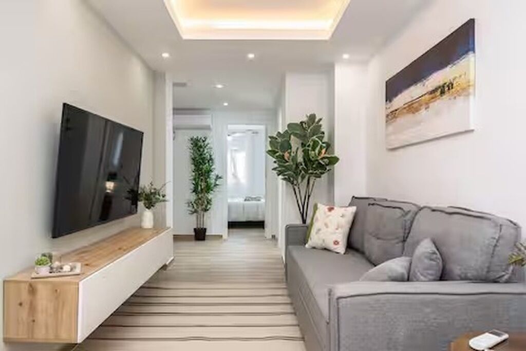 En moderne stue med et fladskærms TV på væggen, en grå sofa med dekorative puder, et træsofabord og en stor stueplante.