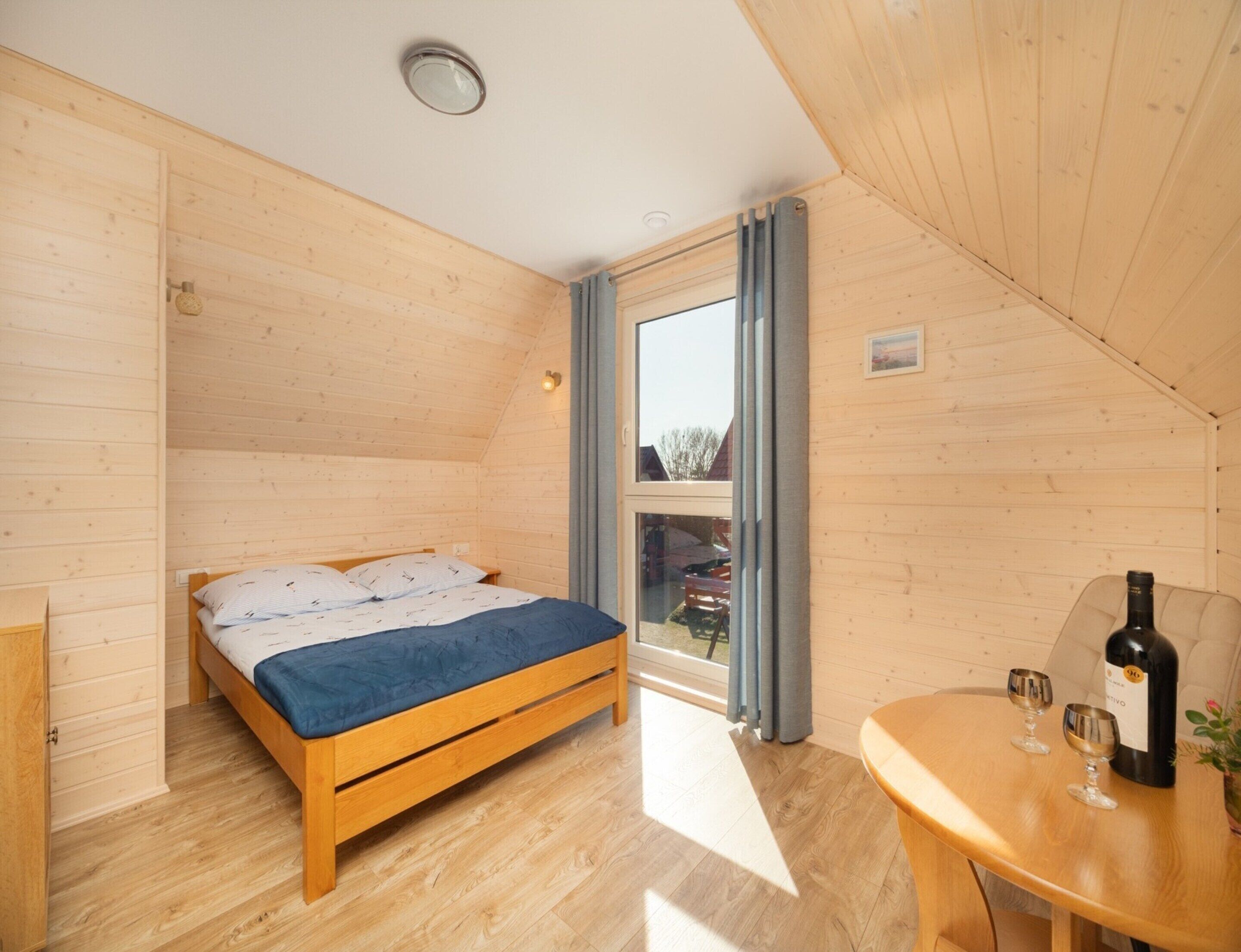 A wooden room with a bed, a table with wine glasses, and a window.