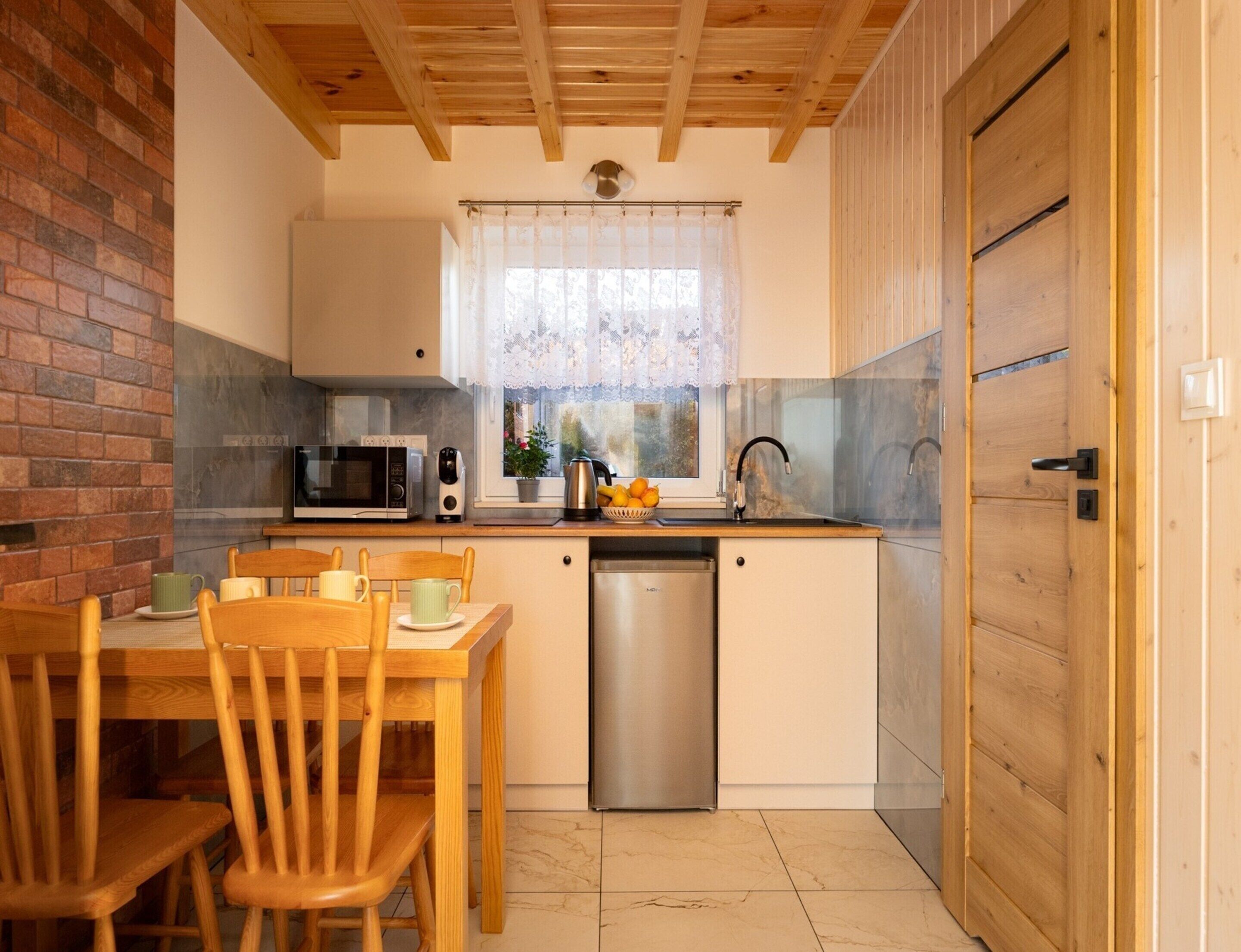 A compact kitchen with a microwave, toaster, and sink. There is a dining table with chairs and a window with lace curtains.