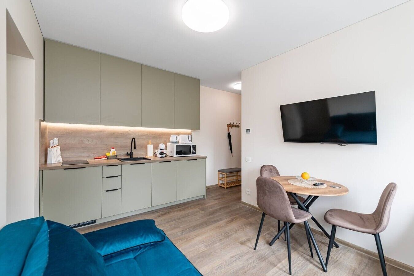 A modern kitchen with a flat-screen TV, grey cabinets, and a dining area with two chairs and a table.