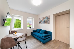 Une pièce avec un canapé bleu, une table à manger avec des chaises, un téléviseur fixé au mur et une porte.