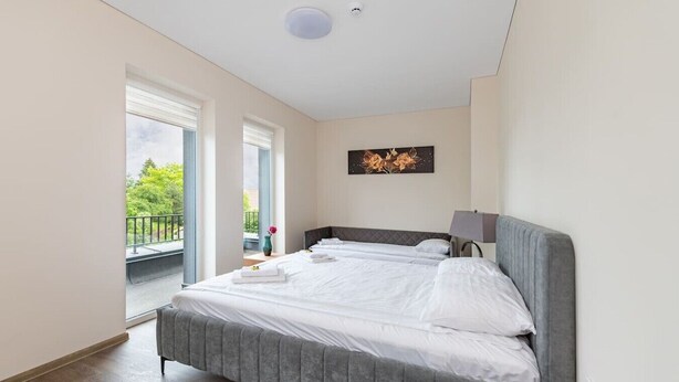 A bedroom with a bed, a nightstand, a lamp, a painting, and a balcony with a railing.