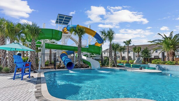 Una piscina con uno scivolo acquatico, palme e un patio in mattoni con sedie blu.