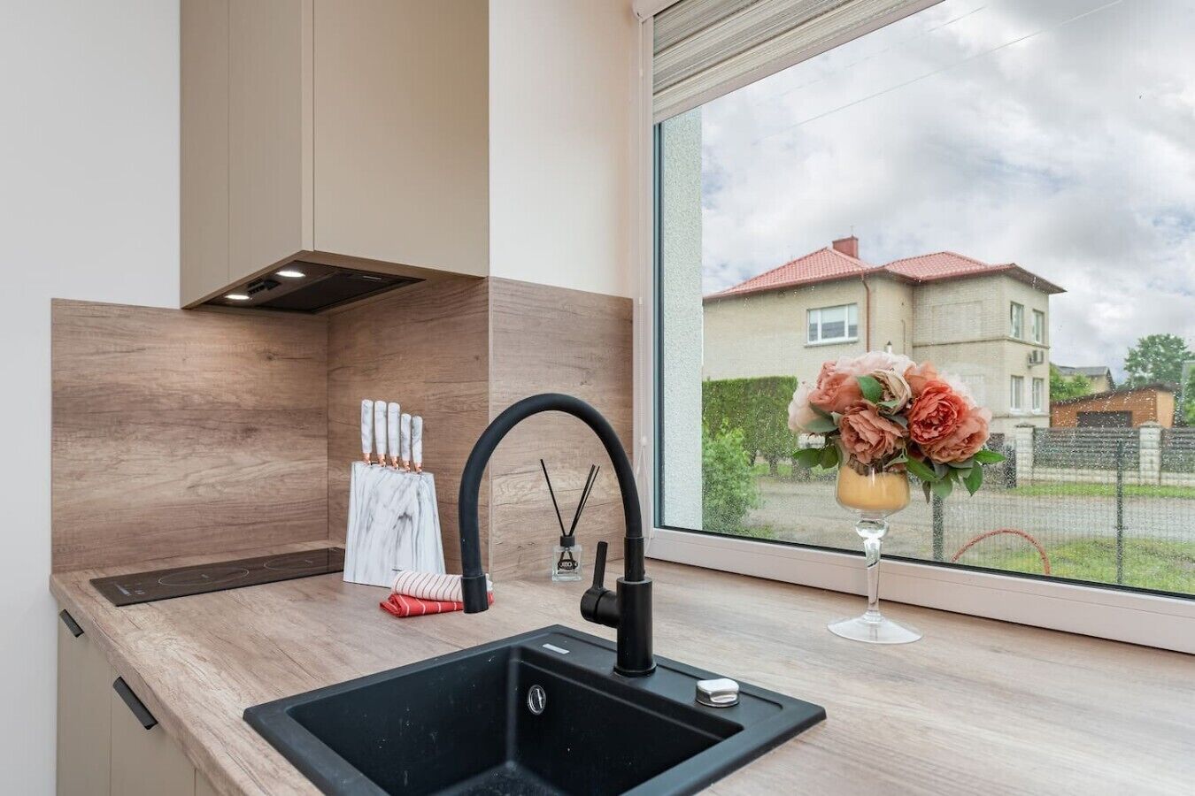 Une cuisine moderne avec un évier noir, une fenêtre donnant sur une maison et un vase rempli de fleurs.