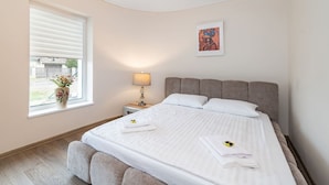 Una camera da letto con un letto, un comodino, una lampada, una finestra con fiori e un quadro appeso al muro.