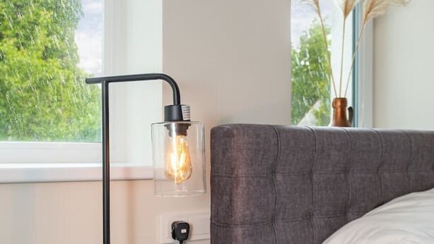 A modern bedroom with a grey upholstered bed, a bedside table with a lamp, and a view of greenery through the window.