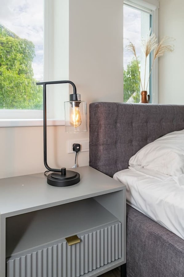 A modern bedroom with a grey upholstered bed, a bedside table with a lamp, and a view of greenery through the window.