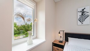A bedroom with a bed, a nightstand, a lamp, a window with a view of greenery, and a wall-mounted artwork.