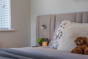 Ein Bett mit Kopfteil, eine Nachttischlampe, ein Teddybär und ein Fenster mit Jalousien.