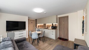 A modern living room with a flat-screen TV, grey sofas, a dining area with a round table and chairs, a kitchen with wooden cabinets and appliances, and a coat rack with an umbrella.