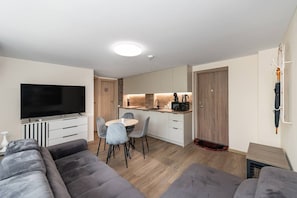 A modern living room with a flat-screen TV, grey sofas, a dining area with a round table and chairs, a kitchen with wooden cabinets and appliances, and a coat rack with an umbrella.