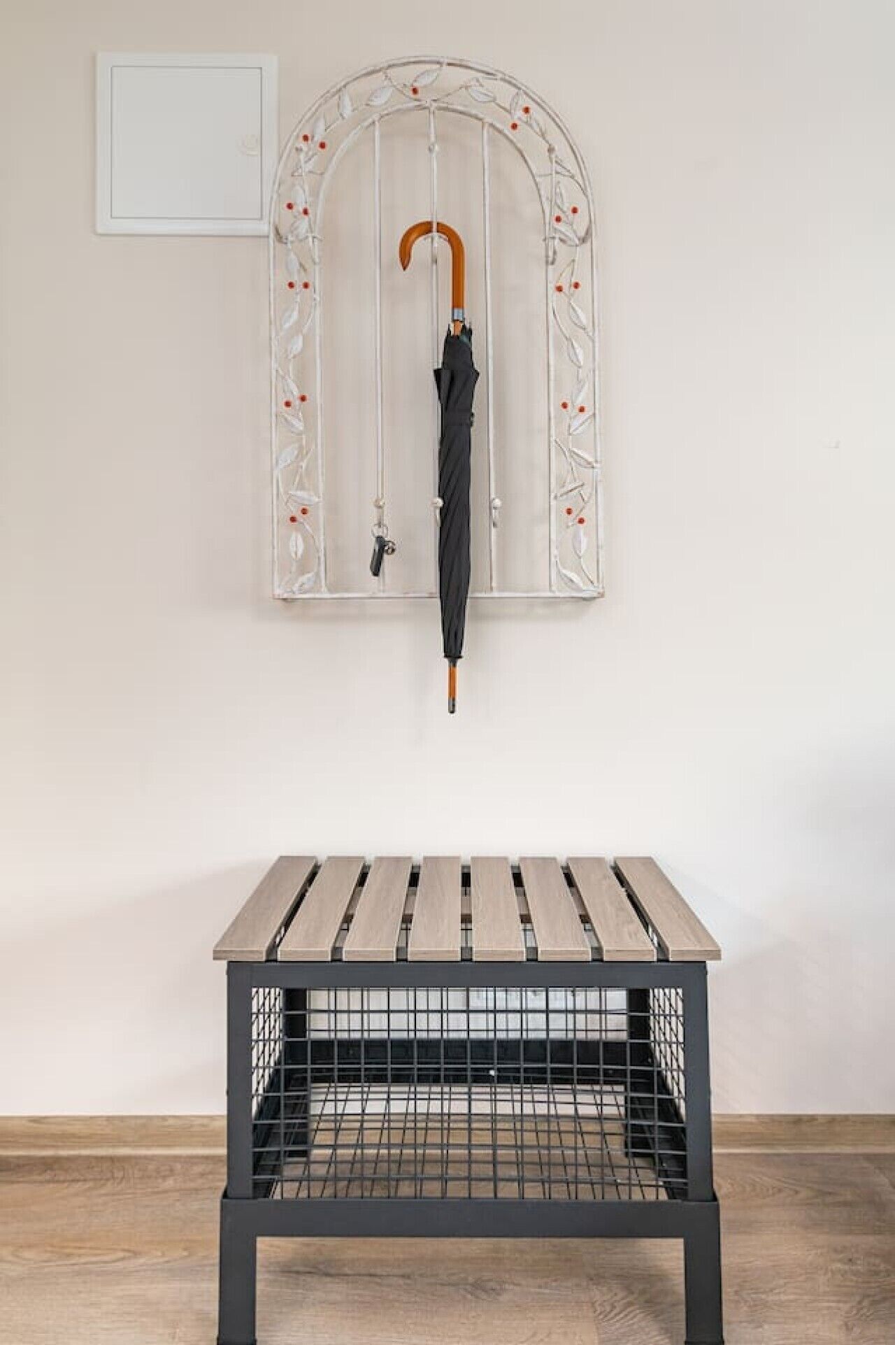 Een tafel met een metalen frame en houten latten, een aan de muur bevestigde vogelkooi met een parasol, en een eenvoudige muur met een klein, ingelijst kunstwerk.