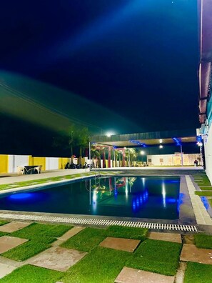 A swimming pool with a surrounding deck, a covered seating area, and a building in the background. - Kanha Green (Bhopal)