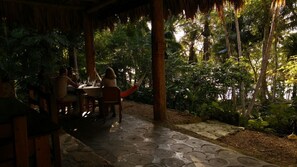 An outdoor dining area with a thatched roof, stone flooring, and lush greenery.