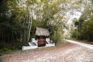 Una estructura rústica con techo de paja identificada como 'El Sombrero Ecological Lodge', con un sendero de tierra que atraviesa un bosque.