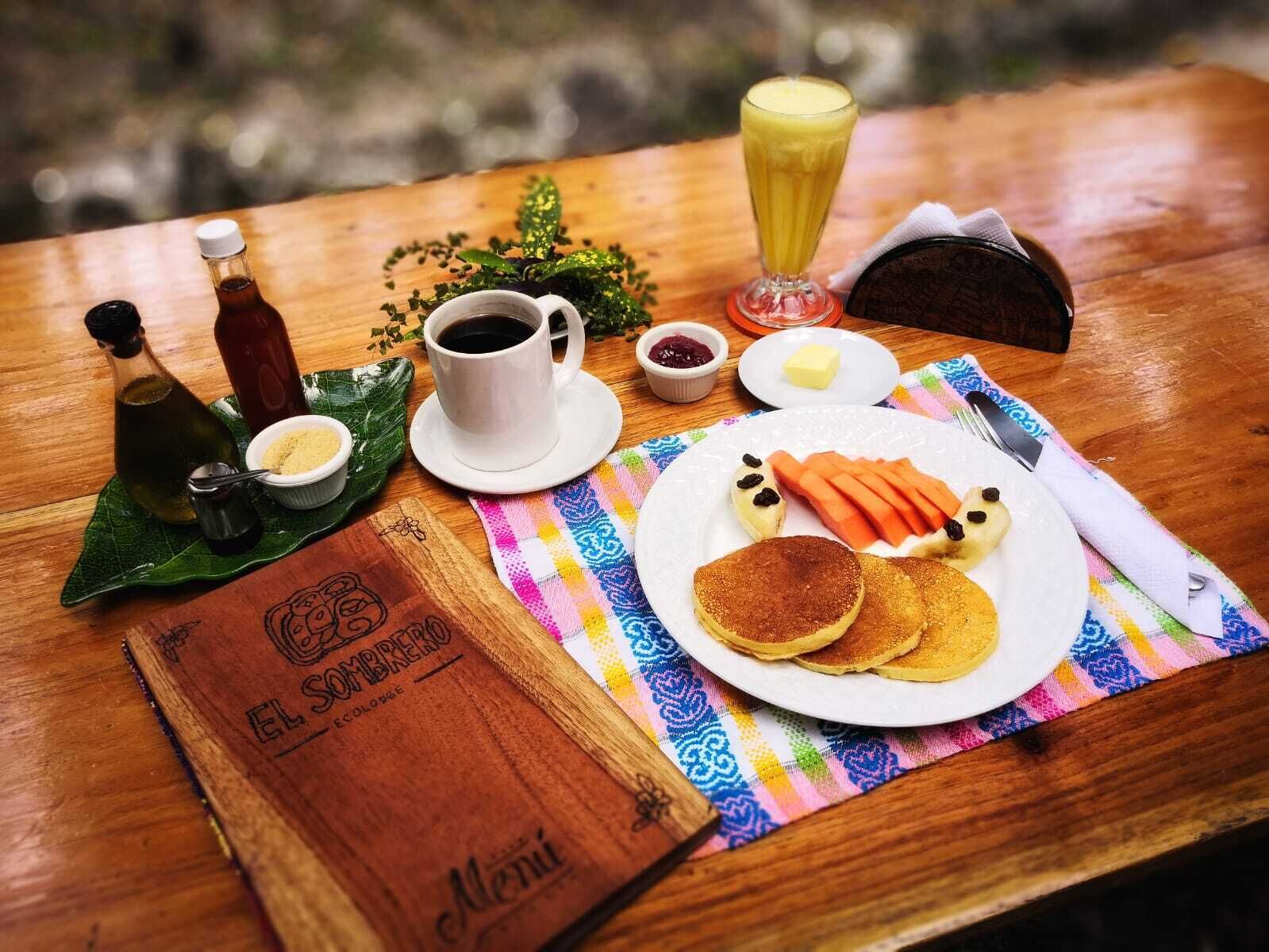 Una mesa de madera con un desayuno que incluye panqueques, una bebida y salsas.