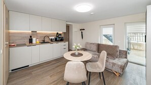 Une cuisine moderne avec des meubles blancs, un coin repas avec une table ronde et deux chaises, un canapé gris et un balcon avec une balustrade.