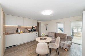 Eine moderne Küche mit weißen Schränken, ein Essbereich mit rundem Tisch und zwei Stühlen, ein graues Sofa und ein Balkon mit Geländer.