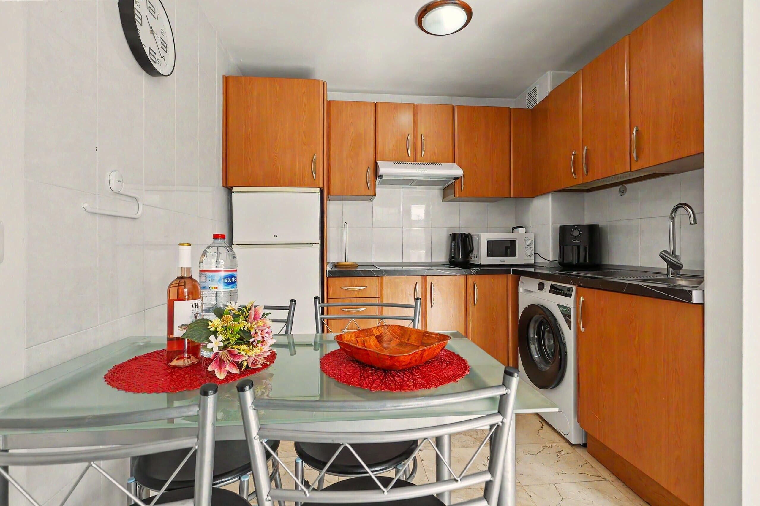 Une cuisine compacte dotée de meubles en bois, d’un four à micro-ondes, d’un lave-linge et d’une table à manger en verre avec des chaises.
