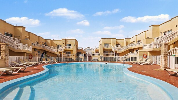 A swimming pool with lounge chairs and a building with balconies. - Vacation Apartment 'Olga' with Shared Pool, Private Terrace and Wi-Fi (Castillo Caleta de Fuste)