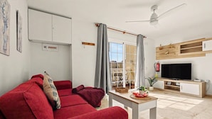 A living room with a red sofa, a flat-screen TV, a ceiling fan, and a window with curtains. - Vacation Apartment 'Olga' with Shared Pool, Private Terrace and Wi-Fi (Castillo Caleta de Fuste)
