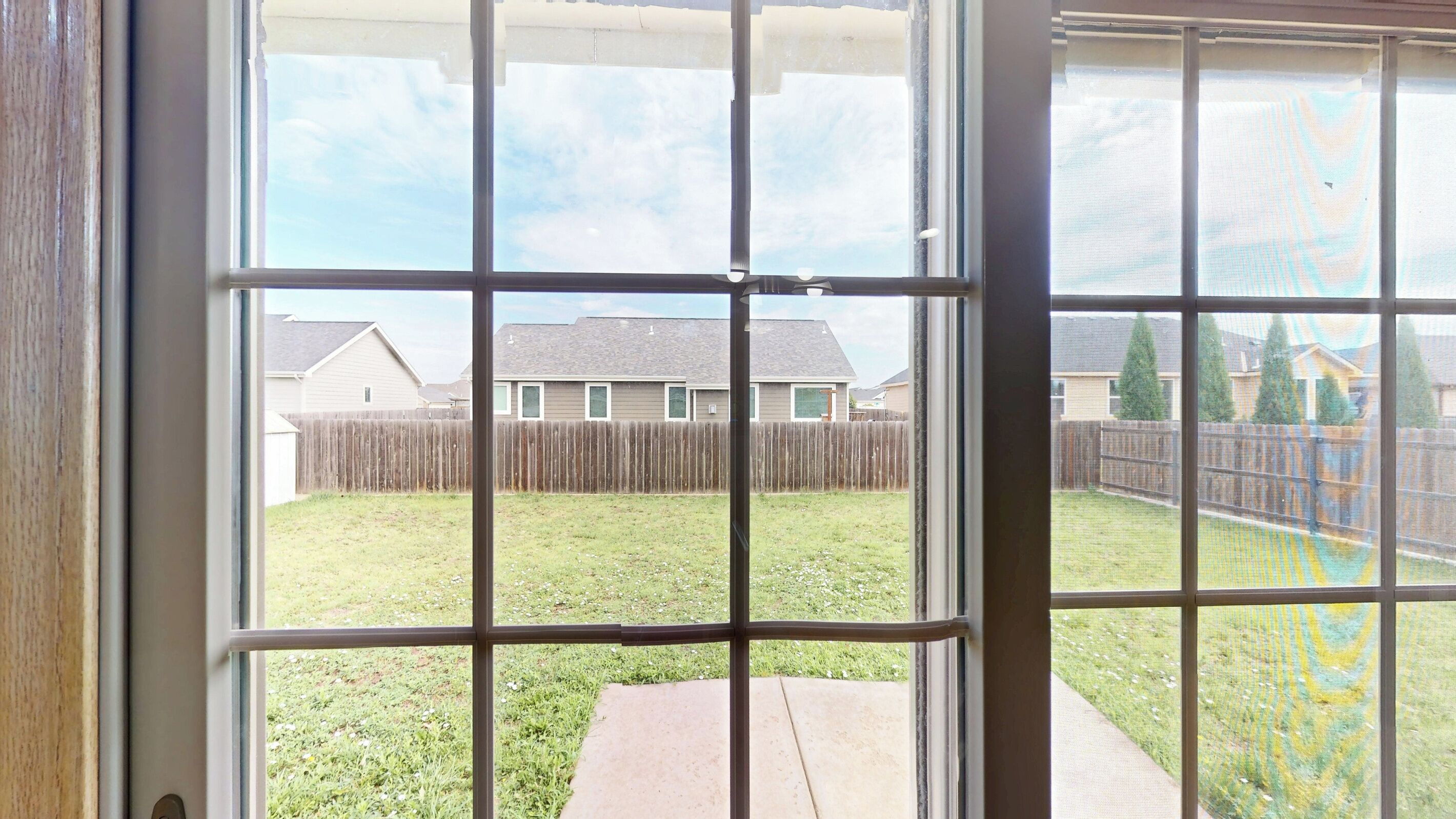 A view through a window with a wooden fence, a grassy yard, and a house in the background.