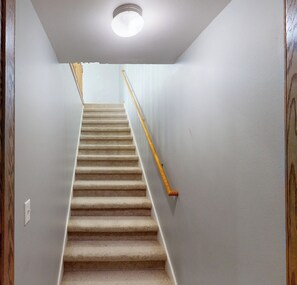 A carpeted staircase with a wooden handrail, white walls, and a ceiling light fixture.