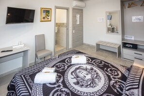 A hotel room with a bed, a desk, a chair, a TV, a mirror, and a shower area. - At the Court of the Gods - Demeter Room (Tropea)