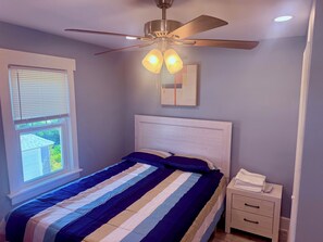 Une chambre à coucher avec un lit, un ventilateur de plafond, une table de chevet et une fenêtre avec des stores.