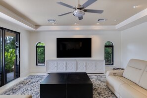 A modern living room with a flat-screen TV, a ceiling fan, and a large window.