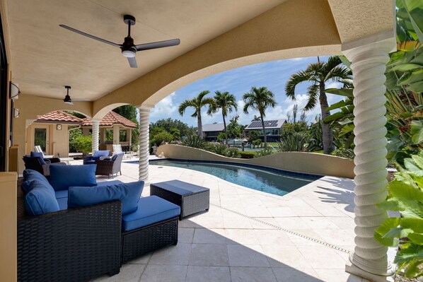 An outdoor pool area with a covered seating area, palm trees, and a building with a solar panel in the background.