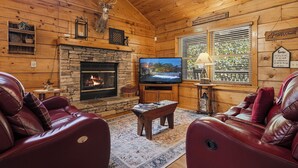 Interior de una cabaña de troncos con chimenea de piedra, sofás de cuero, televisor y ventana con persianas.