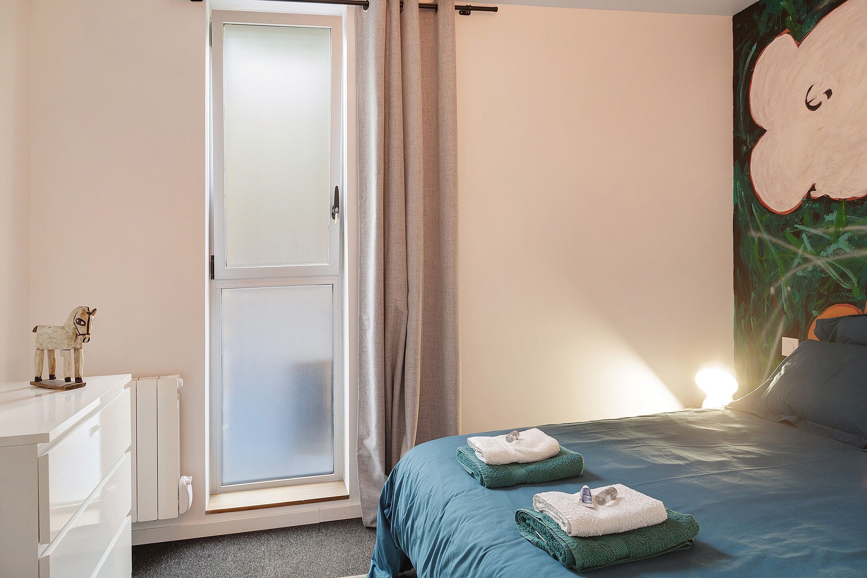 Un dormitorio con cama, un radiador, un aparador blanco, una puerta con cortinas y una pared con un cuadro de nubes.