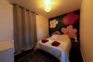 Quarto de hotel com cama, mesa de cabeceira, luminária, parede com obra de arte floral e cortina.