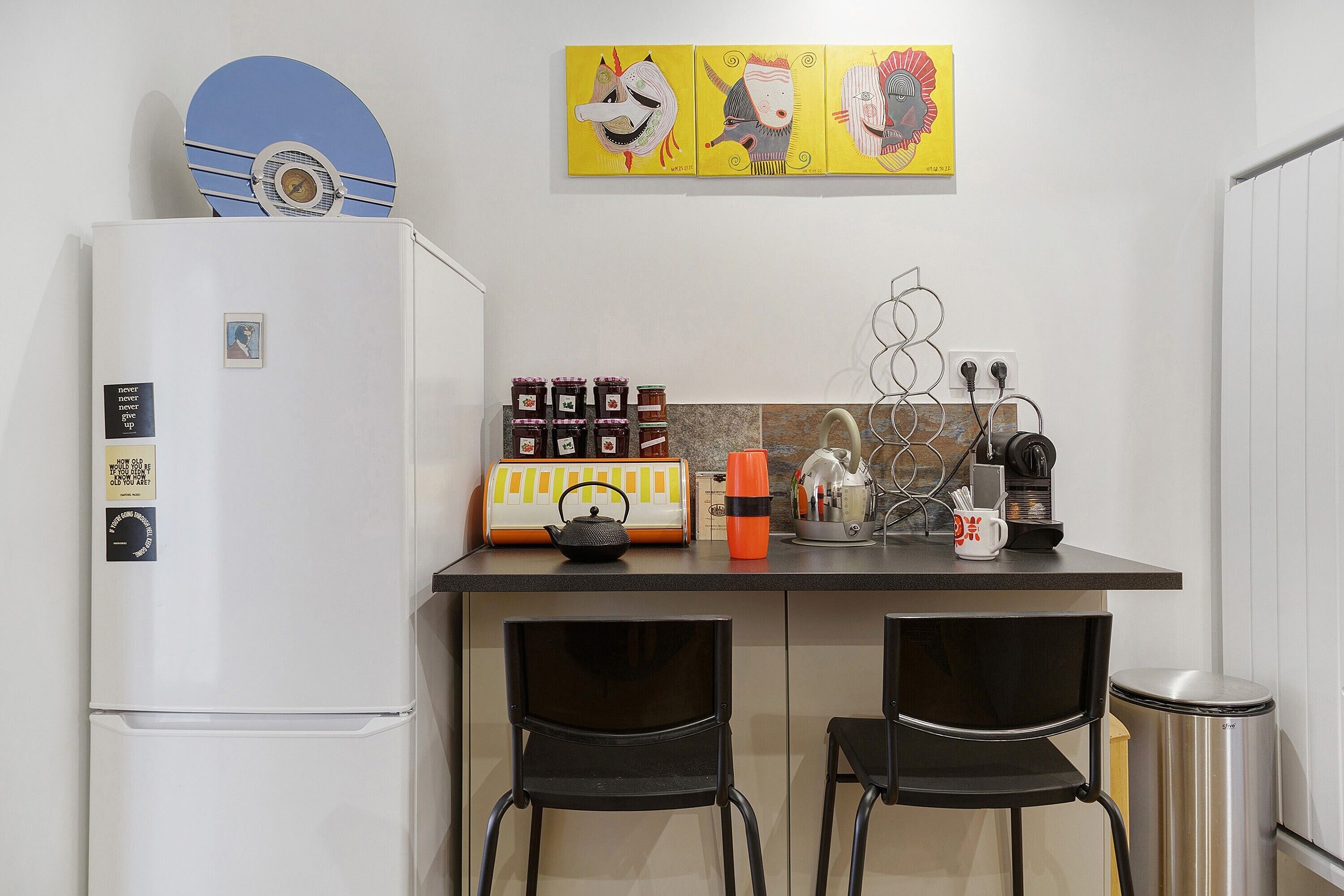 A compact kitchen with a refrigerator, a coffee machine, and a small dining area with two chairs.