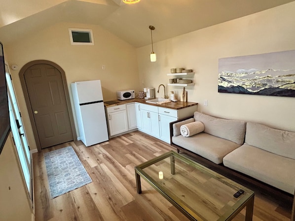 Una cocina compacta con refrigerador, microondas, pileta y encimera de madera. Hay una mesa de centro de vidrio y un sofá color beige con una toalla enrollada.