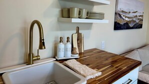 A kitchen with a white sink, gold faucet, and wooden countertop. There are storage shelves above the countertop holding dishes and mugs. - Petite Palace: Guest home in Louisburg, NC (Louisburg)