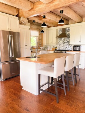 A kitchen with wooden beams, a stainless steel refrigerator, a large island with bar stools, and a patterned backsplash.