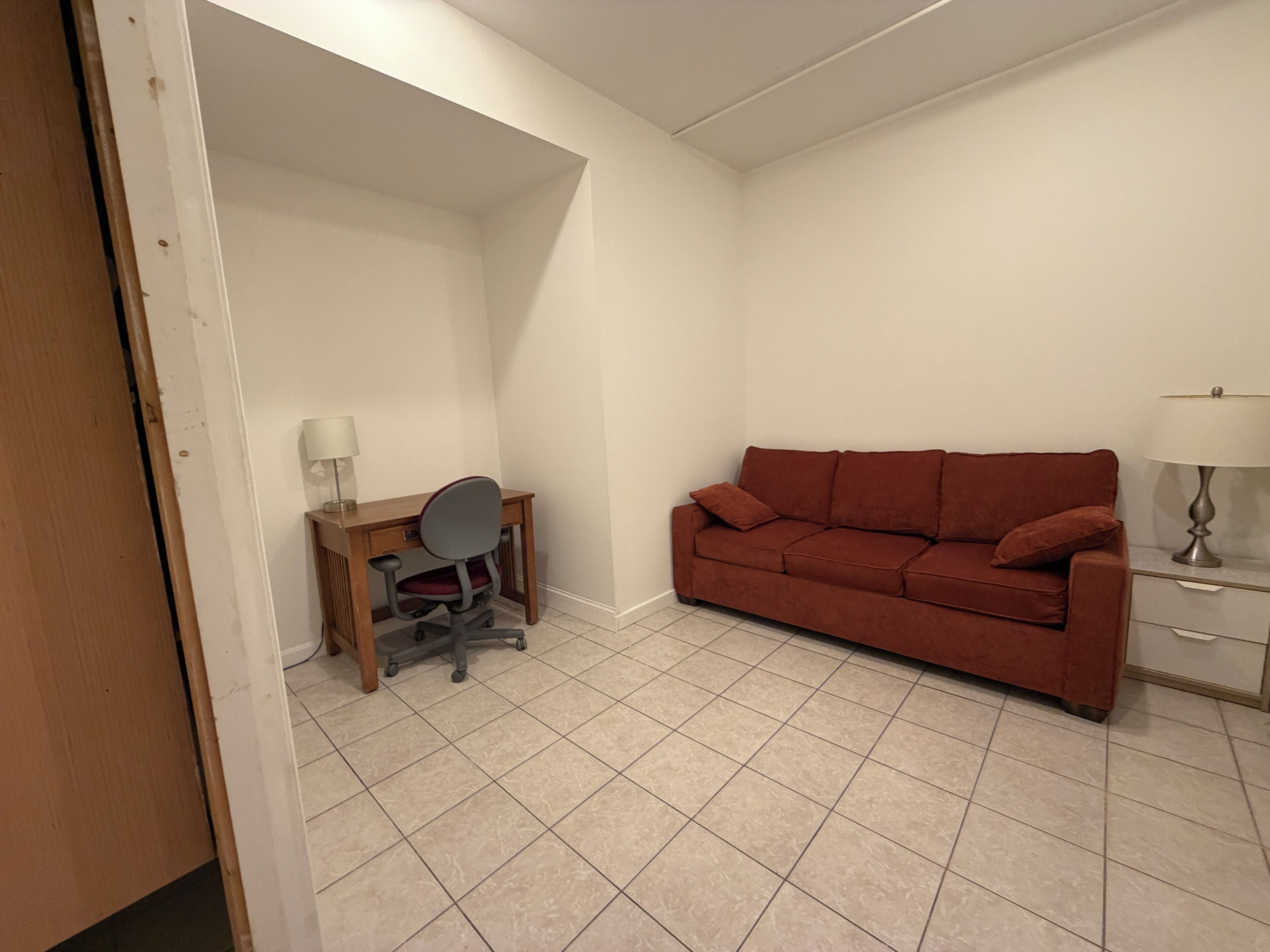 A room with a desk, chair, sofa, and a lamp.