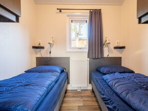 Ein Zimmer mit zwei Betten, einem fensterbelaubten Fenster, einem Heizkörper und Wandlampen.