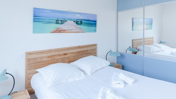 Quarto de hotel moderno com cabeceira de madeira, cama com roupa de cama branca, mesa de cabeceira com abajur, pintura de um cais e um espelho grande que reflete o quarto.
