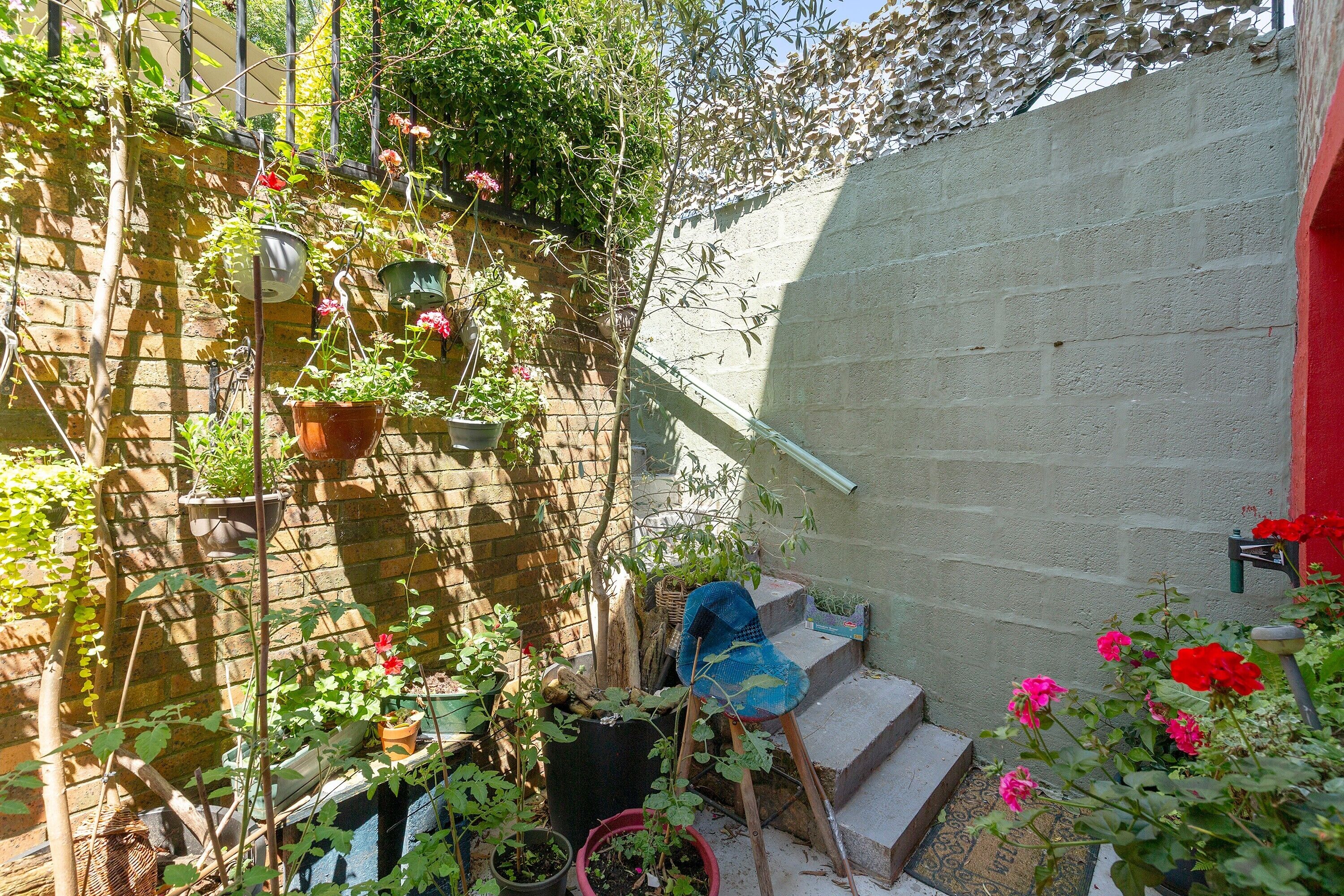 Un petit espace extérieur avec des marches menant à une porte, entouré de plantes en pot et d’une chaise.