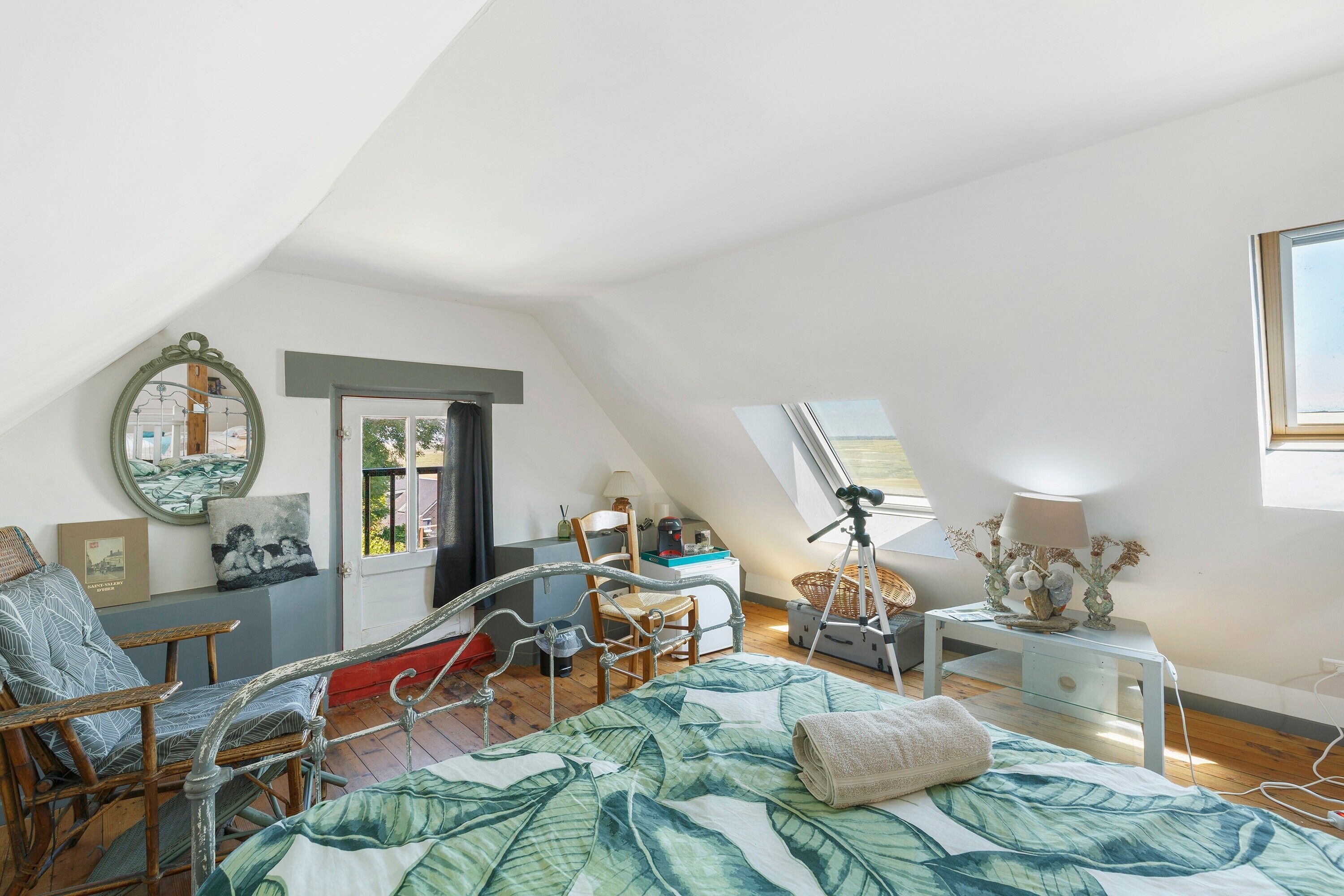 Slaapkamer met schuine kap, een bed met een bedspreide met bladmotief, een stoel, een bureau en een statief.
