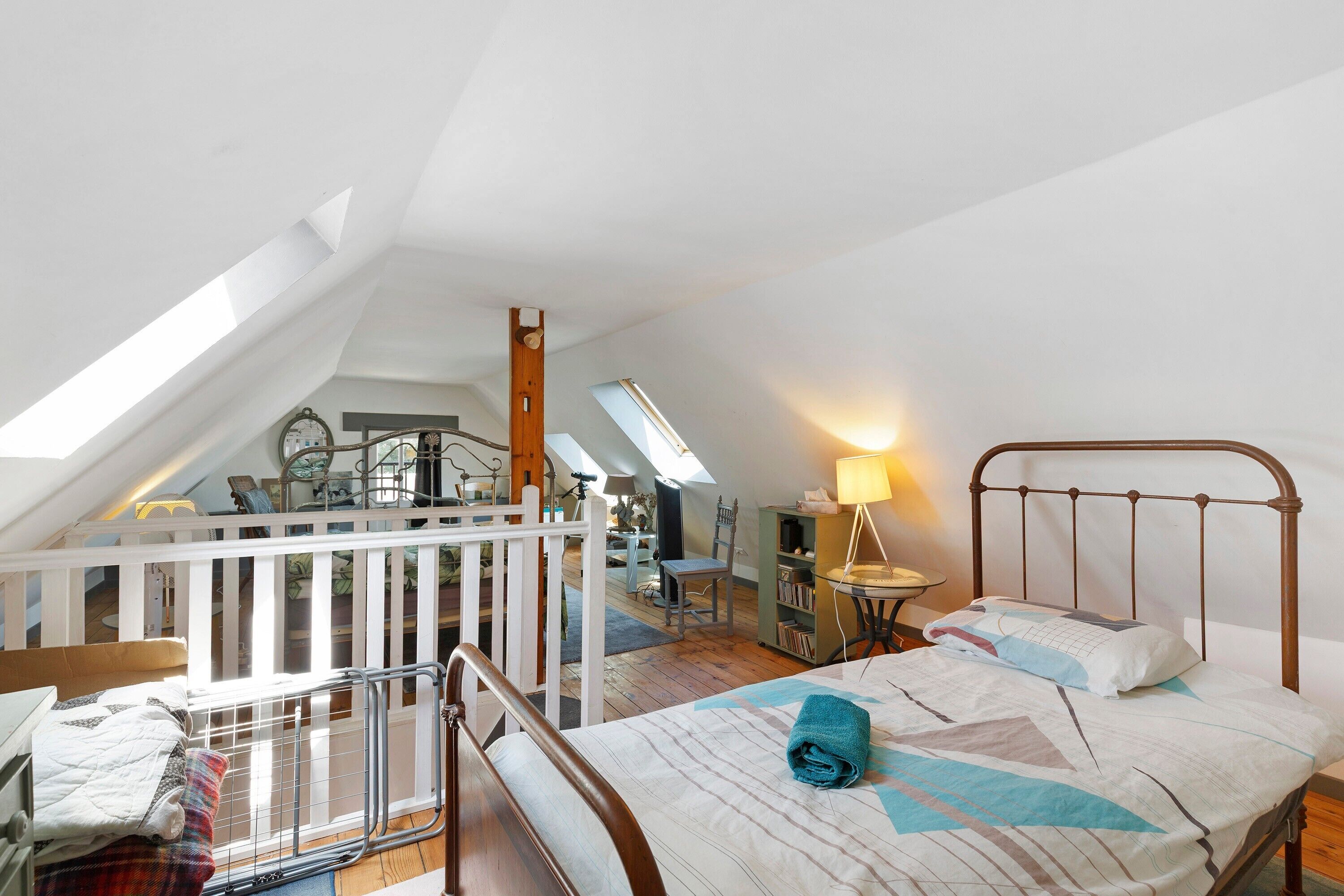 Une chambre à coucher aménagée dans les combles, avec un plafond en pente, un lit avec un cadre en métal, un bureau avec une lampe et une vue sur l’espace de vie.