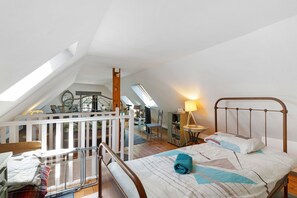 A loft bedroom with a sloped ceiling, a bed with a metal frame, a desk with a lamp, and a view of the living area.