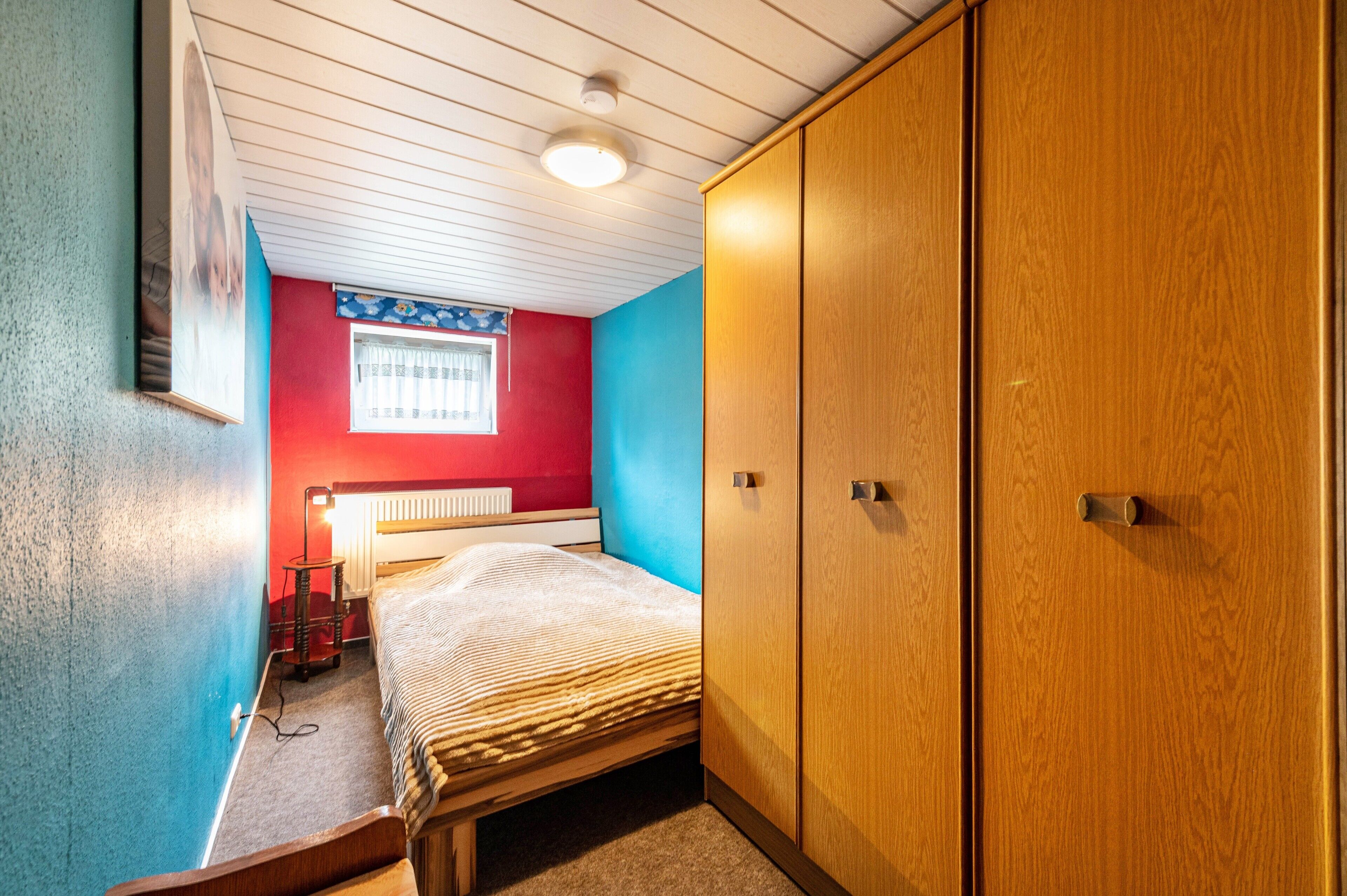 A bedroom with a bed, a radiator, a window with curtains, and a wardrobe.