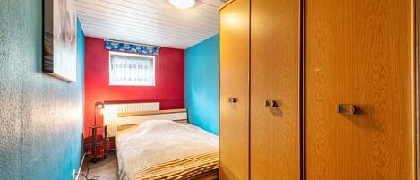 A bedroom with a bed, a radiator, a window with curtains, and a wardrobe.