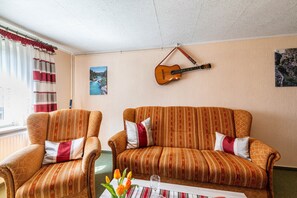 Ein Zimmer mit zwei Polstersesseln, einem Sofa, einer an der Wand hängenden Gitarre und einem fenster mit Vorhängen.